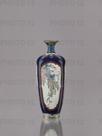 Vase, cloisonné enamel, ovoid elongated form with a short, thin, strongly flared neck. Decoration in four reserves depicting blossoming cherry, apple tree, wisteria, and lilies on a navy blue background. Japanese, ca. 1910–1920 Meiji/Taisho periods. Vessel; height 15.5 cm, diameter 6.0 cm., A tall, narrow vase with a flared rim stands against a neutral gray background, predominantly featuring deep blue and white surfaces with accents of gold, teal, purple, pink, green, and brown; the body displays elongated white panels framed in brown on a dark blue ground, each panel decorated with cascading floral clusters and leafy stems in shades of purple, pink, teal, and green, with delicate outlining and small brown branches; the neck is blue with gold and teal decorative motifs and a gold rim, while the base has decorative bands in blue, teal, and gold; the vase casts a subtle reflection on the surface below.