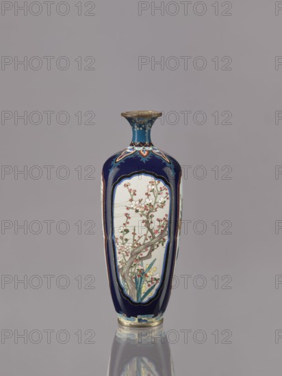 Vase, Ovoid elongated cloisonné enamel vase with a short, thin, strongly flared neck. Decoration: four reserves depicting blossoming cherry, apple tree, wisteria, and lilies on a navy blue background. Japan, ca. 1910–1920 Meiji/Taisho periods. Vessel; height 15.5 cm, diameter 6.0 cm., A tall, narrow vase with a flared rim sits upright against a plain gray background, predominantly colored deep blue with panels of pale off-white featuring a delicate floral design showing branches and blossoms in shades of pink and white with green leaves and brownish-gray stems; the vase also includes accents of gold, turquoise, red, and yellow in decorative borders and small painted details, and the surface reflects faintly below creating a subtle mirrored effect.