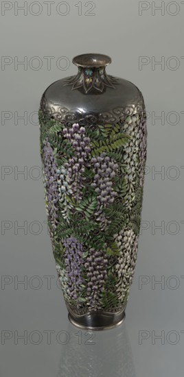 Vase by Hiratsuka Mohei 1836-?, ca. 1880–1890 Meiji period, 1868–1912, Japan. Slender ovoid, double-walled bottle on a very low ring foot with a narrow neck flaring at the spout. Outer wall is openwork composed of densely arranged, colorfully enamelled cloisonné wisteria flowers and leaves filling the vessel's body. Flowers rendered naturalistically in clusters of lilac and blue using multiple enamel shades; leaves in several tones of green. Lower body has a strip of silver left around the foot engraved with a stylized wave motif. The arms of the vessel are solid silver, with a strip of stylized clouds above the floral ornament. Neck is enclosed by a rosette with petals decorated with a multicolored miniature stylized plant ornament featuring chrysanthemum half-rosettes, leafy forms, and volutes. Mark on the bottom: oval convex plaque with the artist's name: Hiratsuka. Vessel; height 19.1 cm, diameter 7.2 cm., A tall, narrow vase with a metallic top and base is covered in raised floral...