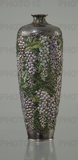 Vase by Hiratsuka Mohei b. 1836 maker, Meiji period, ca. 1880–1890, Japan. Slender ovoid, double-walled bottle on a very low ring foot with a narrow neck flaring at the spout. Outer wall is openwork composed of densely arranged, colorfully enamelled cloisonné wisteria flowers and leaves filling the body. Flowers rendered naturalistically in shades of lilac and blue with multiple enamel tones; leaves in several green tones. Lower body has a strip of exposed silver around the foot engraved with a stylized wave motif. The arms of the vessel are solid silver, with a strip of stylized clouds above the floral ornament. Neck enclosed by a rosette with petals decorated with a multicolored miniature stylized plant ornament including chrysanthemum half-rosettes, leafy forms, and volutes. Mark on the bottom: oval convex plaque with the artist's name: Hiratsuka. Vessel; height 19.1 cm, diameter 7.2 cm., A tall, narrow vase with a rounded shoulder and flared neck sits against a neutral background; ...