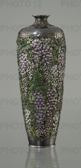 Vase by Hiratsuka Mohei 1836-?, ca. 1880–1890 Meiji period, Japan. Slender ovoid, double-walled bottle on a very low ring foot with a narrow neck flaring at the spout. The outer wall is openwork composed of densely arranged, colorfully enamelled cloisonné wisteria flowers and leaves covering the vessel body. Flowers are rendered naturalistically in shades of lilac and blue using multiple enamel tones; leaves use several green tones. Lower body has a strip of silver around the foot engraved with a stylized wave motif. The arms of the vessel are solid silver with a strip of stylized clouds above the floral ornament. The neck is enclosed by a rosette with petals decorated with a multicolored miniature stylized plant ornament including chrysanthemum half-rosettes, leafy forms, and volutes. Mark on the bottom: oval convex plaque with the artist's name: Hiratsuka. Vessel; height 19.1 cm, diameter 7.2 cm., A tall, narrow vase with a rounded shoulder and flared neck covered in raised floral de...