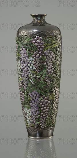 Vase, Hiratsuka, Mohei 1836-? author. A slender ovoid, double-walled bottle on a very low ring foot with a narrow neck flaring at the spout. The outer wall is openwork and composed of densely arranged, colorfully enamelled cloisonné wisteria flowers and leaves, filling the entire surface of the vessel's body. The flowers are rendered naturalistically with clusters in shades of lilac and blue using multiple enamel tones; the leaves employ several green tones. In the lower body, a strip of silver left around the foot is engraved with a stylized wave motif. The arms of the vessel are also solid silver, with a strip of stylized clouds running above the floral ornament in a manner characteristic of Far Eastern art. The neck is enclosed by a rosette with petals decorated with a multicolored miniature stylized plant ornament including chrysanthemum half-rosettes, leafy forms, and volutes. Mark on the bottom: oval convex plaque with the artist's name: Hiratsuka. Ca. 1880–1890 Meiji Period, 186...