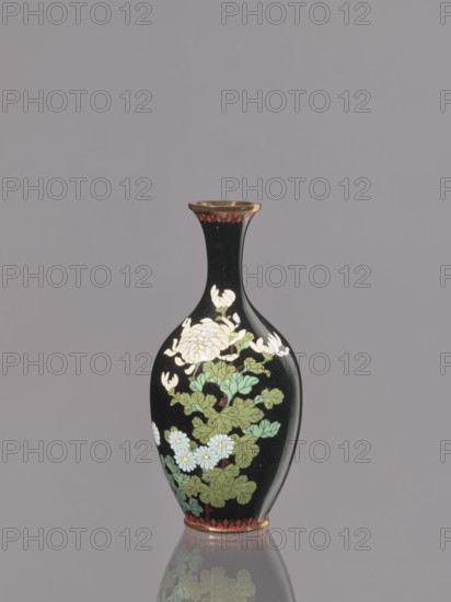 Vase, cloisonné enamel; symmetrical decoration of chrysanthemums and flowers; Meiji period, ca. 1900; Nagoya, Japan; vessel; height 19.3 cm, diameter 9.1 cm., A slender vase with a narrow neck and flared rim sits against a plain gray background, the vase surface predominantly black with decorative floral motifs in white, green, pale blue, and touches of red and gold; the floral design features a group of petals and leaves wrapping around the body, and a band of red decoration encircles the rim and base, with the vase reflected faintly on the surface beneath it.