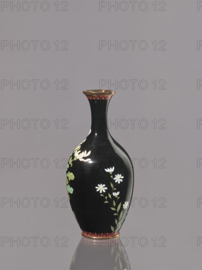 Vase, chrysanthemums, Japanese art, Japanese culture, flowers iconography, Meiji 1868–1912, cloisonné enamel, symmetrical decoration, ca. 1900, Nagoya, Japan, vessel; height 19.3 cm, diameter 9.1 cm., A glossy black vase with a narrow neck and flared rim stands against a muted gray background, decorated with painted white flowers and green leaves along its rounded body, a band of red decorative scallops near the rim and base, and a reflective surface that creates a subtle mirrored reflection below.