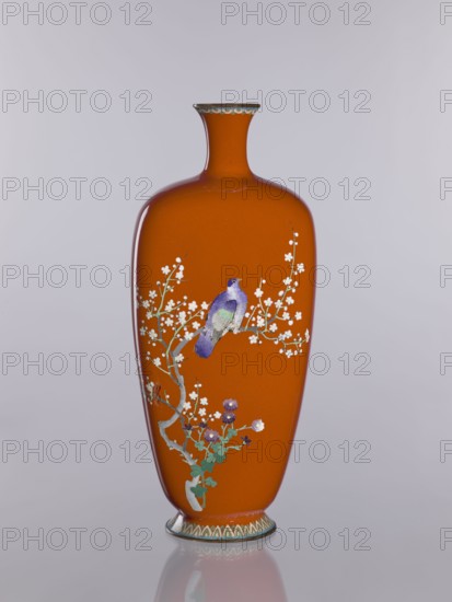 Vase, cloisonné enamel; decorated with chrysanthemums, cherries, and birds; Meiji–Taisho period, ca. 1910–1920; Nagoya, Japan; height 24.0 cm, width 10.4 cm, depth 8.5 cm., An orange vase with a narrow neck and flared rim stands against a pale gray background, decorated with a painted branch bearing small white blossoms and green leaves; a purple and blue bird perches on the branch, and a few purple flowers appear near the base, while the rim and foot have subtle patterned bands in muted green and cream.