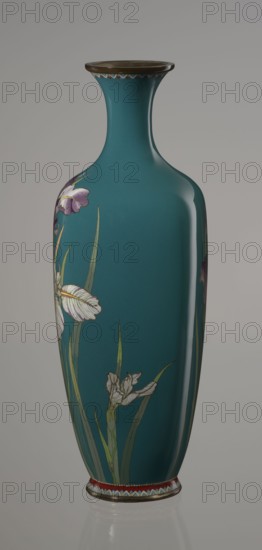 Vase, cloisonné enamel, ovoid vase one of a pair, slender, on a low ring-shaped foot and with a narrow neck, flaring at the rim. The foot and mouth are framed with sheet metal. Decorated with a motif of a blooming iris bush, captured with naturalistic fidelity and attention to a variety of color tones. Executed using the yusen technique, the motif is placed against a uniform sea-green background. Around the foot and the rim there are narrow, colorful borders, also made using the yusen technique, composed of small triangular forms called cicada wings. Ca. 1900 Meiji period, 1868–1912, Japan, Nagoya. Vessel; height 25 cm, width 9.7 cm, depth 8.8 cm., A tall vase with a narrow neck and flared rim sits against a neutral background; the vase is predominantly teal and has painted floral motifs featuring a few long green leaves and several flowers with white and pale purple petals, with hints of yellow and darker purple in some petals; the rim and base have metallic brown edges and a decorati...