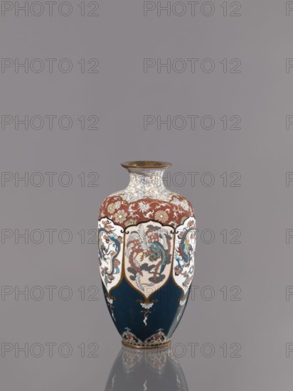 Vase, cloisonné enamel; Meiji period ca. 1890–1900. Vase on a small ring-shaped foot with a short neck ending in a spout widening toward the top. Decoration: stylized plant ornament and alternating dragon and phoenix ho-o motifs in six reserves; Japanese dragon and phoenix iconography. Kyoto, Japan. Vessel. Height 15.0 cm; diameter 8.0 cm., A ceramic vase with a narrow neck and flared rim sits against a plain gray background, showing a palette of deep blue, red, white, gold, teal, brown, and touches of black; the lower body is predominantly deep blue with gold and white decorative motifs, the midsection features white cartouche-like panels each containing a multicolored stylized bird and floral designs in teal, red, brown, and blue, separated by gold outlines, and the upper shoulder displays a red field with repeating floral patterns in gold and white, while the neck has intricate blue and white scrolling designs and the rim appears metallic brown, with the vase casting a faint reflect...