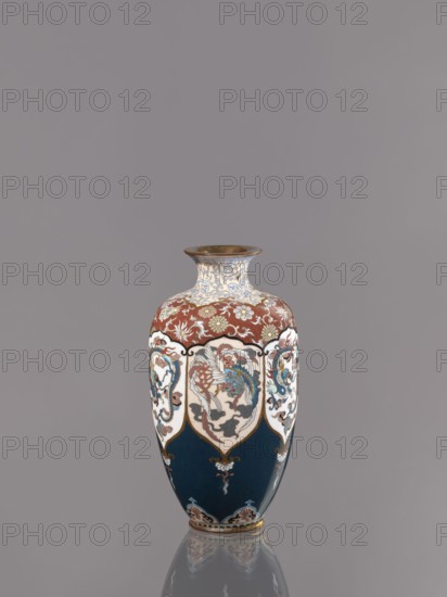 Vase, cloisonné enamel. Vase on a small, ring-shaped foot, with a short neck ending in a spout widening towards the top. Decoration: stylized plant ornament, alternating dragon and phoenix in six reserves Japanese dragon motif, hoo/phoenix, ca. 1890–1900 Meiji period, 1868–1912, Japan, Kyoto. Vessel; height 15.0 cm, diameter 8.0 cm., A decorative vase stands against a plain grey background, showing a glossy surface with visible reflection beneath it. The vase displays a palette of blue, deep teal, white, red, gold, brown, and hints of cream. Its upper neck area has intricate floral and scroll patterns in cream and gold on a white background, transitioning to a red band with floral motifs. The midsection features shield-shaped white panels containing stylized bird and cloud-like designs in blue, red, gold, and brown, separated by gold outlines. The lower portion is deep teal with small ornamental flourishes in gold and white and a decorative base rim in gold and cream. The overall shape...