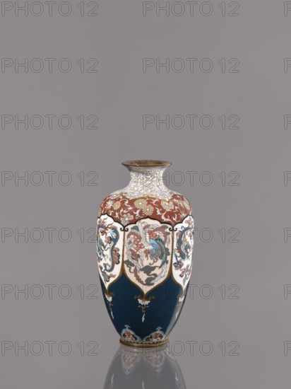 Vase, Cloisonné enamel. Vase on a small ring-shaped foot with a short neck ending in a spout widening toward the top. Decoration: stylized plant ornament, alternating dragon and phoenix in six reserves. Japanese dragon motif, hoo phoenix, dragons, plant ornament. Meiji period, ca. 1890–1900. Japan, Kyoto. Vessel. Height 15.0 cm, diameter 8.0 cm., A decorative vase stands against a plain gray background, with a glossy surface reflecting faintly on the base. The vase has a flared rim with a metallic brown edge and a narrow neck patterned in pale cream with blue and gold scrolling motifs. Below the neck, a band of deep reddish-brown with gold floral ornament wraps around the shoulder. The main body features a repeating shield-like panel design on a white ground, each panel containing ornate stylized floral and feathered motifs in shades of blue, red, cream, and brown accented with gold outlines. The lower portion of the vase is a deep navy blue with small decorative gold and cream element...