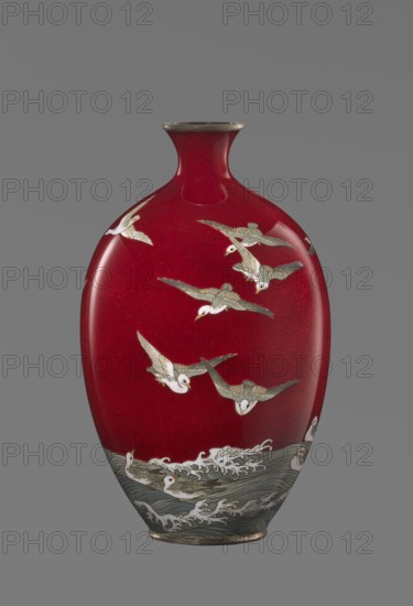 Vase by Hayakawa Komejirô fl. ca. 1880–1912. Small ovoid, bulging bottle on a very low round foot, framed in silver-plated metal, with a short narrow neck and flared spout also framed in silver-plated metal. Body covered with transparent red enamel akasuke technique, developed around 1880 in Nagoya. Motif of water birds—plovers Japanese: chidori—executed with the yusen technique using thin silver wires, depicted flying over foaming waves or perching on the water, treated with a combination of naturalism and decorative stylization typical of Japanese design. Brand signature engraved in kanji on the bottom: Hayakawa zo tsukuru, the mark of enameller Hayakawa Komejirô. Late 19th century Meiji period, 1868–1912. Origin: Nagoya, Japan. Vessel; height 16.7 cm, diameter 9.9 cm., An oval-shaped vase with a narrow neck and flared rim sits against a plain gray background; the vase surface is a deep red with a glossy finish and is decorated with a group of pale birds in flight rendered in white a...