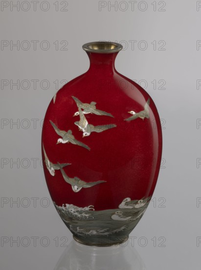 Vase by Hayakawa Komejirô fl. ca. 1880–1912. Small ovoid, bulging bottle on a very low round foot framed in silver-plated metal, with a short narrow neck and flared spout also framed in silver-plated metal. Body covered with transparent red enamel akasuke technique, developed around 1880 in Nagoya decorated with a motif of water birds—plovers chidori—executed using the yusen technique with thin silver wires, shown flying over foaming waves or perching on the water. Motif combines naturalism and decorative stylization typical of Japanese design. Brand signature engraved in kanji on the bottom: Hayakawa zo tsukuru, the enameller’s mark. Late 19th century Meiji period, 1868–1912, Nagoya, Japan. Vessel. Height 16.7 cm; diameter 9.9 cm., A glossy vase with a narrow neck and rounded body sits against a neutral background; the main color of the vase is deep red with a metallic rim at the mouth, and it is decorated with a group of flying birds rendered in pale gray and white with darker gray a...