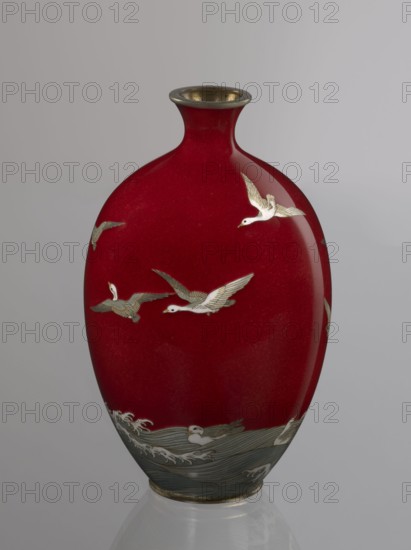 Vase by Hayakawa Komejirô fl. ca. 1880–1912. A small ovoid, bulging bottle on a very low round foot framed in silver-plated metal, with a short narrow neck and flared spout also framed in silver-plated metal. The body is covered with transparent red enamel akasuke technique, developed around 1880 in Nagoya, decorated with a motif of water birds—plovers chidori—rendered using the yusen technique with thin silver wires, shown flying over foaming waves or perching on the water. The design combines naturalism and decorative stylization characteristic of Japanese design. The maker’s signature is engraved in kanji on the bottom: Hayakawa zo tsukuru. Late 19th century Meiji period, 1868–1912, Nagoya, Japan. Vessel height 16.7 cm, diameter 9.9 cm., A glossy vase with a deep red body and a rounded, slightly elongated shape topped by a narrow neck with a metallic rim; the vase is decorated with a group of white birds in flight with pale gray and gold detailing on their wings and bodies, and a st...