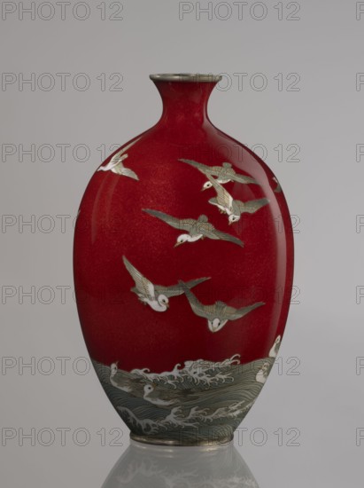 Vase by Hayakawa Komejirô fl. ca. 1880–1912. Small ovoid, bulging bottle on a very low round foot framed in silver-plated metal, with a short narrow neck and flared spout also framed in silver-plated metal. Body covered with transparent red enamel akasuke technique, developed around 1880 in Nagoya, decorated with a motif of water birds — plovers chidori — rendered with the yusen technique using thin silver wires, shown flying over foaming waves or perching on the water. Motif combines naturalism and decorative stylization typical of Japanese design. Signature engraved in kanji on the bottom: Hayakawa zo tsukuru, the mark used by enameller Hayakawa Komejirô. Late 19th century Meiji period, 1868–1912, Nagoya, Japan. Vessel; height 16.7 cm, diameter 9.9 cm., A smooth vase with a glossy deep red surface decorated with a group of flying birds and a scene of waves and swimming birds near the base; the bird figures are rendered in pale white and muted gray-green with fine dark outlines, and t...