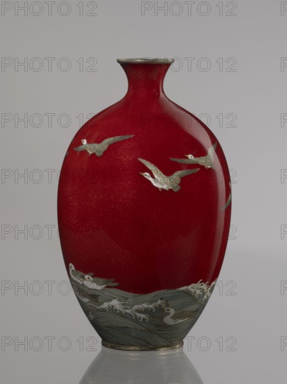 Vase by Hayakawa Komejirô fl. ca. 1880–1912. Small ovoid, bulging bottle on a very low round foot framed in silver-plated metal, with a short narrow neck and flared spout also framed in silver-plated metal. Body covered with transparent red enamel akasuke technique, developed ca. 1880 in Nagoya decorated with water birds—plovers chidori—rendered with the yusen technique using thin silver wires, shown flying over foaming waves or perching on the water. Motif combines naturalism and decorative stylization typical of Japanese design. Signature engraved in kanji on the bottom: Hayakawa zo tsukuru, the mark used by enameller Hayakawa Komejirô. Late 19th century Meiji period, 1868–1912, Nagoya, Japan. Vessel; height 16.7 cm, diameter 9.9 cm., A smooth, glossy vase with a deep red body and a narrow neck topped by a metallic rim, decorated with a group of pale birds in flight across the upper area and a few pale birds floating on stylized grey-blue waves at the lower section, with accents of w...