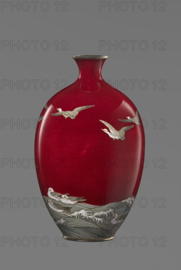 Vase, Hayakawa, Komejirô fl. ca. 1880–1912? craftsman. A small ovoid, bulging bottle on a very low round foot framed in silver-plated metal, with a short narrow neck and flared spout also framed in silver-plated metal. The body is covered with transparent red enamel akasuke technique, developed around 1880 in Nagoya, decorated with a motif of water birds—plovers Japanese: chidori—executed with the yusen technique using thin silver wires, shown flying over foaming waves or perching on the water. The motif combines naturalism and decorative stylization typical of Japanese design. The maker’s mark is engraved in kanji on the bottom: Hayakawa zo tsukuru, the signature used by enameller Hayakawa Komejiro. Late 19th century Meiji period, 1868–1912, Nagoya, Japan. Vessel; height 16.7 cm, diameter 9.9 cm., A smooth vase with a deep red glossy surface set against a neutral gray background, shaped with a narrow neck and a slightly flared rim that appears metallic. A group of pale birds with gray...
