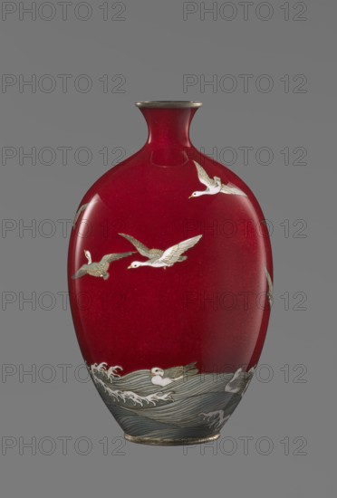 Vase by Hayakawa Komejirô fl. ca. 1880–1912, late 19th century Meiji period, 1868–1912, Nagoya, Japan. Small ovoid, bulging bottle on a very low round foot framed in silver-plated metal, with a short narrow neck and flared spout also framed in silver-plated metal. Body covered with transparent red enamel akasuke technique, developed around 1880 in Nagoya. Motif of water birds—plovers chidori—rendered with the yusen technique using thin silver wires, depicted flying over foaming waves or perching on the water, combining naturalism and decorative stylization typical of Japanese design. Signature engraved in kanji on the bottom: Hayakawa zo tsukuru. Vessel; height 16.7 cm, diameter 9.9 cm., A glossy vase with a deep red body and a narrow neck topped by a metallic rim, decorated with a few white and gray birds in flight and a group of birds floating on stylized gray and white waves near the base; the scene includes metallic-looking accents on the birds and rim, and the background is a flat...