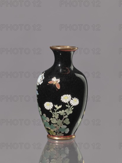 Vase with a short, flared neck, ca. 1890–1910 Meiji period, 1868–1912, Japan, Nagoya; cloisonné enamel; height 12.1 cm, diameter 6.8 cm. Decoration: chrysanthemums, butterflies, flowers iconography., A glossy vase with a narrow neck and flared rim sits against a plain background; the vase surface is predominantly black with a few small reflective highlights and areas of speckling, and it features painted floral motifs including white daisy-like flowers with yellow centers, green leaves in varying shades, and a butterfly with orange and white markings. The rim and base have metallic gold tones with a thin band of muted red near the base, and the vase casts a faint reflection on the surface beneath it.