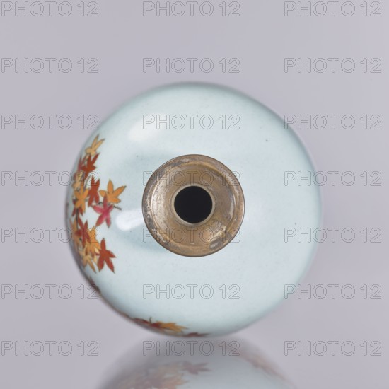 Vase, cloisonné enamel, ovoid elongated form with a narrow spout, decorated with chrysanthemums and maples plant ornaments/flowers iconography. Japanese, Nagoya; Meiji–Taisho period, ca. 1910–1920. Height 12.4 cm; diameter 16.3 cm., A round ceramic object viewed from above with a glossy pale blue surface, a central circular metal rim that is brass-colored and shows wear, and a cluster of small leaf-shaped decorations in shades of red, orange, and yellow along one side, all set against a soft grey background with a faint reflection of the object.