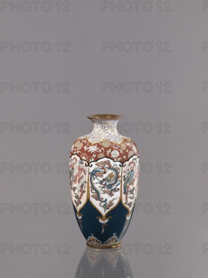 Vase, cloisonné enamel. Vase on a small, ring-shaped foot with a short neck ending in a spout widening toward the top. Decoration: stylized plant ornament in six reserves alternating dragon and phoenix motifs. Japanese dragon and phoenix hoo iconography. Meiji period, ca. 1890–1900. Japan, Kyoto. Vessel; height 15.0 cm, diameter 8.0 cm., A tall ceramic vase with a narrow neck and flared rim standing on a reflective surface, decorated with intricate painted panels and floral motifs. The vase shows a palette of deep blue, rich red, white, gold, light blue, green, and brown. The upper portion features red background areas with white floral designs and gold accents, while the main body alternates white shield-shaped panels containing stylized dragon- or floral-like figures outlined in gold against deep blue sections with decorative gold elements. The neck is primarily white with delicate light blue and gold scrolling patterns, and the rim and base have metallic gold finishes. The overall b...