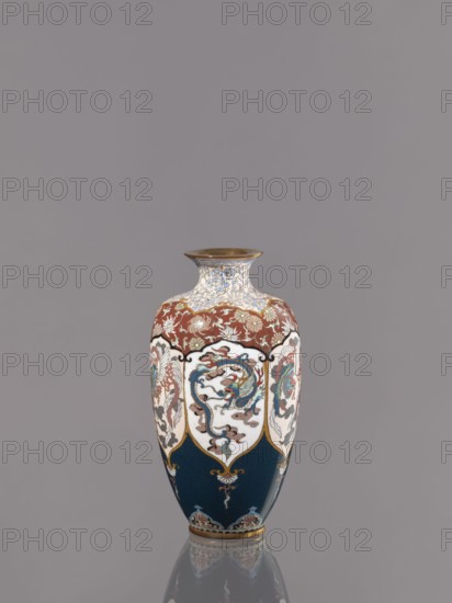 Vase, gift; repossession Kraków. Cloisonné enamel vase on a small ring-shaped foot with a short neck ending in a spout widening toward the top. Decoration: stylized plant ornament in six reserves alternating dragon and phoenix Japanese dragon motif, hoo/phoenix, Meiji period, ca. 1890–1900. Japan, Kyoto. Vessel. Height 15.0 cm, diameter 8.0 cm., A single vase sits centered against a plain gray background, with a rounded body that tapers to a narrow neck and flared rim; the vase displays a palette of deep blue, white, red, gold, and touches of light blue and green, with ornate painted panels featuring flowing, curving motifs and stylized creatures, floral patterns along the shoulder in red and gold, decorative gold outlines separating the panels, and a reflective surface that creates a faint mirrored reflection on the surface below.