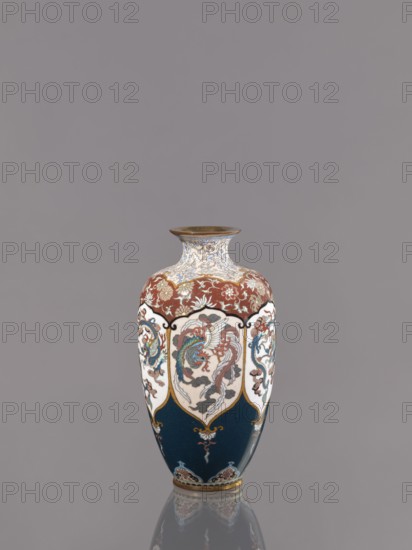 Vase, Cloisonné enamel, Meiji period ca. 1890–1900, Kyoto, Japan. Vase on a small ring-shaped foot with a short neck ending in a spout widening toward the top; decorated with stylized plant ornament in six reserves alternating dragon and phoenix Japanese dragon motif, hoo/phoenix. Height 15.0 cm; diameter 8.0 cm., A single decorative vase sits centered against a plain gray background, with its reflection faintly visible below; the vase has a narrow neck flaring to a small rim with a metallic-looking top edge, a bulbous shoulder with ornate floral and scroll patterns in shades of red, white, gold, blue, and pale beige, and a lower body that transitions to a deep navy blue with gold accents and intricate multicolored motifs including stylized bird-like figures and cloud-like shapes, with additional detailing in black and muted green.