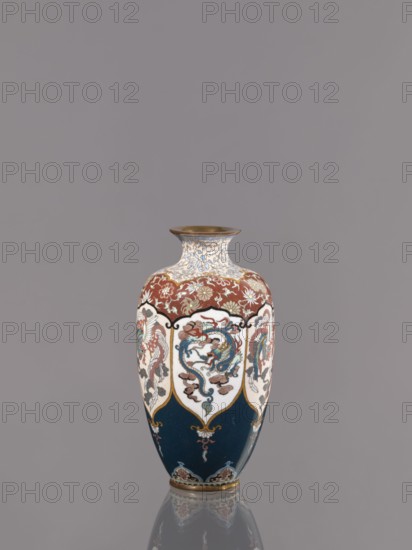 Vase by, Meiji period ca. 1890–1900, Kyoto, Japan. Cloisonné enamel vessel on a small ring-shaped foot, with a short neck ending in a spout widening toward the top. Decoration: stylized plant ornament in six reserves alternating dragon and phoenix Japanese dragon motif and hoo/phoenix. Height 15.0 cm; diameter 8.0 cm., A tall decorative vase sits against a plain gray background, with a narrow neck flaring to a slightly wider rim in a metallic brown tone and a rounded body that tapers to a small base. The vase shows a combination of deep blue, rich red, creamy white, gold, and touches of teal and brown in its painted panels and patterns. The upper shoulder area has a red field with floral motifs in gold and white, while the main body features several white cartouche-like panels each containing a stylized creature rendered in blue, red, teal, and brown, separated by gold outlines. The lower portion of the vase is a deep blue with gold ornamental accents and small decorative motifs near t...