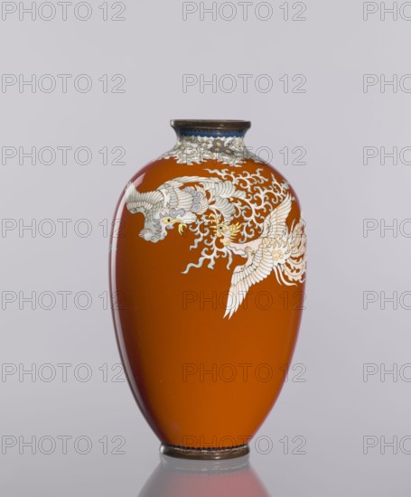 Vase, cloisonné enamel vessel with phoenixes and plant ornaments on a dark brick-red background; Japanese, Meiji–Taisho period, ca. 1910–1920; height 12.8 cm, diameter 7.7 cm., An orange vase with a glossy finish sits against a pale gray background, decorated with a flowing white and pale blue stylized bird motif and swirling cloudlike patterns near its rounded shoulder, a narrow dark rim at the top with a band of blue and light decorative detailing beneath it, and a dark base that reflects slightly on the surface below; visible colors include orange, white, pale blue, dark brown or black, and hints of yellow.