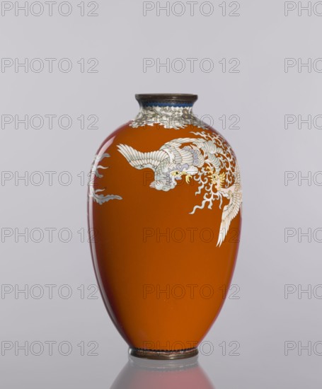 Vase, cloisonné enamel, ca. 1910–1920 Meiji/Taisho periods, Japan. Cylindrical vessel, height 12.8 cm, diameter 7.7 cm. Decoration: phoenixes and plant ornaments on a dark brick-red background. Japanese, An orange vase with a rounded body and narrow neck stands against a pale gray background, decorated with a flowing white and pale blue bird motif with yellow accents and stylized white cloudlike patterns around the neck, a dark rim at the mouth and base, and a subtle reflection beneath it.