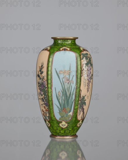 Vase, cloisonné enamel, floral pattern with butterflies and dense green floral rosettes in reserved background. Japanese, Meiji period 1868–1912, 19th/20th century. Vessel; height 15.3 cm, width 8.7 cm, depth 8.2 cm., A tall vase with a rounded body and narrow neck sits against a plain gray background, predominantly featuring green, pale blue, cream, purple, white, and brown tones; the vase has a bright green patterned band around the neck and base, panels of pale blue and cream on the body decorated with floral scenes including long green leaves and pale flowers, clusters of purple and white blossoms, and additional small floral motifs, with metallic-looking rims at the top and bottom and a mirrored reflection visible beneath the vase.