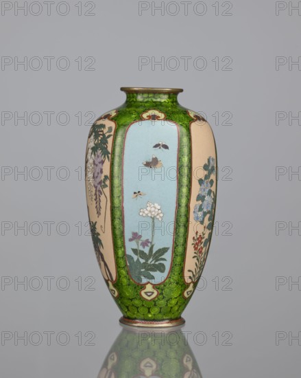 Vase, cloisonné enamel, floral pattern with butterflies and dense green floral rosettes in reserves; Japanese, Meiji period 1868–1912, 19th/20th century; vessel; height 15.3 cm, width 8.7 cm, depth 8.2 cm., A tall vase with a rounded body and narrow neck sits against a plain gray background, predominantly featuring a bright green mottled pattern interspersed with several vertical panels in pale blue and beige that contain botanical and insect motifs; the panels show stylized flowers in colors including white, purple, blue, red, and green, along with a few small flying insects in muted brown and yellow tones, and the vase has thin metallic rims at the top and base with a subtle reflection of the vase visible on the surface below.