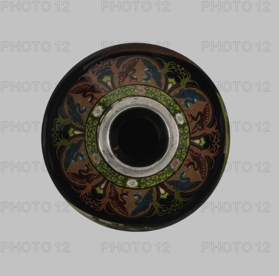 Vase, Namikawa Yasuyuki 1845–1927, enameller. Meiji period ca. 1890–1905, Kyoto, Japan. Small slender cloisonné vase, height 18.0 cm, diameter 7.8 cm. Body tapers to a ring-shaped silver foot and short neck with spout enclosed by a silver ring. Ornamental shippo cloisonné border with rosettes in rhomboid fields at the foot and neck. Petal decoration with stylized phoenixes flowing down to the shoulders. Belly covered with shiny black enamel and six colorful fan-shaped reserves depicting floral and bird motifs executed in the yusen technique with silver wires. Silver plaque mounted in the bottom mirror engraved “Kyoto Namikawa.”, A round vessel seen from above with a central circular opening surrounded by a metallic ring, decorated with an ornate band featuring floral motifs and swirling leaf-like patterns in green, brown, blue, red, and gold on a predominantly black background, with small white flower shapes interspersed among the decoration.