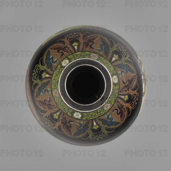 Vase by Namikawa Yasuyuki 1845–1927, cloisonné enamel, ca. 1890–1905 Meiji period. A small, slender vase with a body tapering toward the bottom and terminating in a ring-shaped silver foot. The arms merge into a short neck slightly parted at the spout, enclosed by a silver ring. At the foot is an ornamental shippo motif border with rosettes inscribed in rhomboid fields; a similar ornament adorns the neck. From the neck a decoration of petals filled with stylized phoenix images flows down to the shoulders. The belly is covered with glossy black enamel decorated with six colorful reserves shaped like unfolded fans, each with various floral and bird motifs executed with meticulous precision in the yusen technique using silver wires. On the foot, in the mirror of the bottom, a silver plaque with an engraved signature reads “Kyoto Namikawa.” Kyoto, Japan. Height 18.0 cm; diameter 7.8 cm., A rounded vessel viewed from above with a central circular opening surrounded by a narrow metallic rim ...