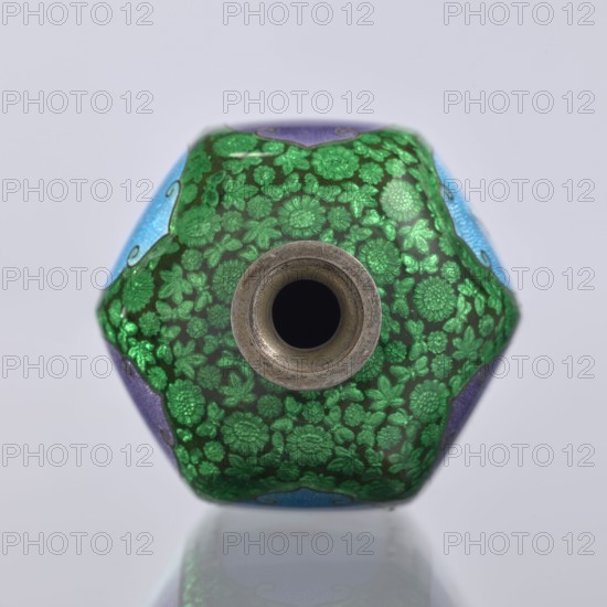 Vase, gift, repossession; elongated hexagonal cloisonné enamel vase with a narrow spout. Decoration: small, densely spaced floral rosettes, irises, butterflies and birds on a black background. Japanese, Meiji period 1868–1912, 19th/20th century. Vessel. Height 12.5 cm; width 6.7 cm; depth 6.1 cm., A close-up view of a small, hexagonal object with a central circular opening; the surface shows a dense floral pattern in bright green on a darker background, with visible sections of blue and purple around the sides, and a metallic rim around the opening that appears silver-gray; the background is pale gray and the object casts a subtle reflection beneath it.