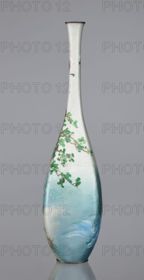 Vase by Kawaguchi Bunzaemon 1860–post 1910, cloisonné enamel, Meiji period, late 19th century. Slender vessel with a long thin neck, height 30.6 cm, width 7.8 cm. Decoration: two pine trees growing on a seaside rock, the rolling sea and two flying cranes. Japan, Nagoya., A tall, narrow vase with a long neck and rounded body stands against a plain gray background; the surface shows a gradient of colors from pale off-white near the neck to light blue and turquoise toward the base, with visible texture and subtle iridescent sheen; a branch with green foliage wraps partway around the upper body, the branch rendered in brown and the leaves in shades of green; the rim and base have thin dark edges in a muted gray or black.