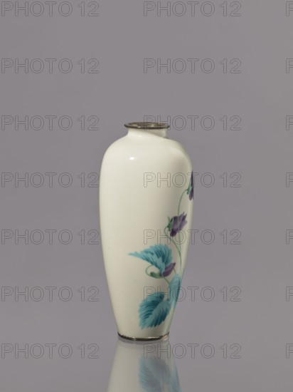 Vase, Flowers iconography. Japanese, Taisho period ca. 1912–1926. Cloisonné enamel. Nagoya, Japan. Vessel with narrow rim; decoration: a tuft of blooming lilac bellflowers along almost the entire height of the belly. Height 15.1 cm; width 6.6 cm., A smooth, cream-colored ceramic vase with a narrow neck and a thin metallic rim stands upright against a muted gray background, decorated on one side with a floral design featuring several teal leaves and a few purple flowers connected by thin green stems, and a soft reflection of the vase appears below on the reflective surface.