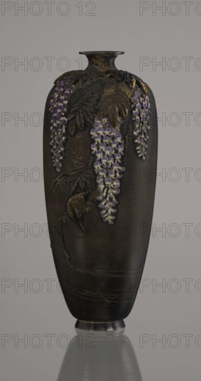 Vase, Toshitsugu maker, Meiji period 19th/20th century, Japan. Ovoid vase on a low ring foot with a very short neck and a strongly flaring, plate-like spout. Decorated in a naturalistic, asymmetrical composition showing a wisteria vine with three clusters of flowers hanging from the top near the neck; a kingfisher perches on a branch near the lowest flower, gazing into engraved water with swimming fish. Decoration executed with ro-iro zogan inlaid color painting combining inlaid metals and alloys with colored enamels white, several shades of lilac, subtle green accents. Leaves and bird figurine are gilded and patinated with shakudo. Lower belly bears engraved inscription: Sumisuiin Toshitsugu sei. Cloisonné enamel vessel, height 16.2 cm, diameter 7.1 cm., A tall dark vase with a narrow neck and flared rim stands against a neutral background, decorated with raised botanical motifs including a few cascading clusters of purple and pale lilac flowers, several elongated leaves rendered in s...