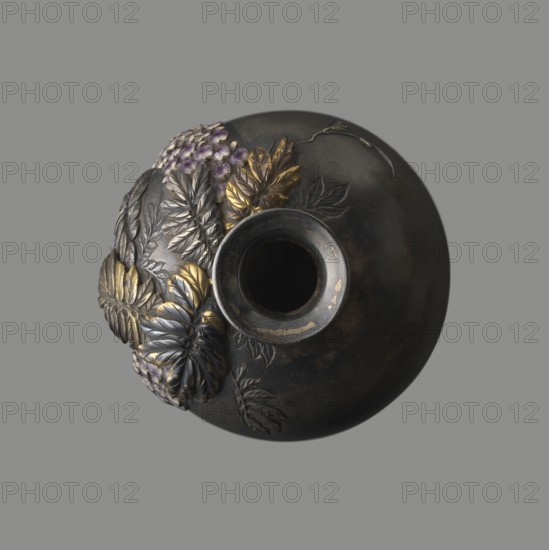 Vase, Toshitsugu author, Meiji period 19th/20th century, Japan. Ovoid vase on a low ring foot with a very short neck and strongly flaring, plate-like spout. Decorated in relief, applied, inlaid and engraved naturalistic motifs arranged asymmetrically: a wisteria vine with three clusters of hanging flowers near the neck; a kingfisher perching on a branch near the lowest flower, gazing into engraved water with engraved fish. Decoration executed in the ro-iro zogan technique inlaid color painting, inlaying various metals and alloys and using colored enamels—white, several shades of lilac, and subtle green accents. Leaves and bird figurine gilded and patinated with shakudo. Lower belly engraved with inscription: Sumisuiin Toshitsugu sei. Vessel, height 16.2 cm, diameter 7.1 cm., A round dark object with a central circular opening sits against a gray background; one side of the object is decorated with a cluster of raised leaves and flower petals in metallic gold and silver tones with touch...