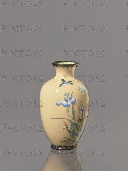 Vase, ; flowers and birds decoration; Meiji period Japan, ca. 1900; cloisonné enamel on vessel; height 12.2 cm, diameter 7.1 cm., A small ceramic vase with a rounded body and narrow flared neck sits against a plain gray background, displaying a creamy yellow surface decorated with painted botanical motifs and a small bird. The decoration includes a group of iris-like flowers with soft blue and pale purple petals and green leaves, plus additional floral elements in pale pink and white, and slender green foliage extending upward and downward. A small bird with blue and dark gray markings appears near the neck. The rim and base have a darker brownish band with scalloped teal accents, and the vase casts a soft reflection on the surface beneath it. The visible colors include gray, creamy yellow, blue, green, pale purple, pink, white, brown, teal, and dark gray.