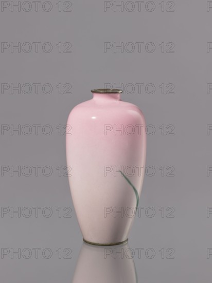 Vase, Taisho period ca. 1912–1926, Japan — Nagoya. Cloisonné enamel vessel decorated with flowers and irises. Height 15 cm; diameter 7.9 cm., A smooth ceramic vase with a rounded shoulder and narrow neck sitting on a reflective surface, primarily pink fading to a lighter pink toward the base, with a small curved green brushstroke on one side; the neck and base edges have a metallic rim in a muted brass color, and the background is a uniform gray.