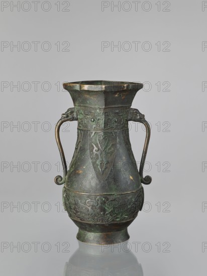 Hu-type vase, hu vessel, Chinese, Ming dynasty 1368–1644, Xuande period 1426–1435, possibly backdated signatures; materials and form: ceramic vessel, ceremonial/ritual hu with taotie ornamentation; dimensions: height 31.5 cm, width 17.0 cm., A single metal vessel with a dark green and brown patina stands against a plain light gray background, featuring a flared octagonal rim, a tapered body with embossed decorative panels, and two curved handles ending in small spirals attached near the upper section; the surface shows areas of darker brown and verdigris green, with intricate raised patterns and a rounded lower body on a slightly flared foot.