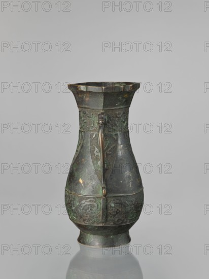Hu-type vase, hu vessel, Chinese, Ming dynasty 1368–1644, Xuande period 1426–1435 attributed, backdated signatures, taotie ornamentation, ceremonial/ritual vessel, height 31.5 cm, width 17.0 cm, China., An aged metal vessel with a narrow neck flaring into a scalloped rim and a rounded body standing on a short foot, featuring a single vertical handle attached from rim to midbody; the surface shows decorative raised motifs in bands around the neck and lower body and worn areas with patina and spots of green and brown, and the background is a smooth gray.