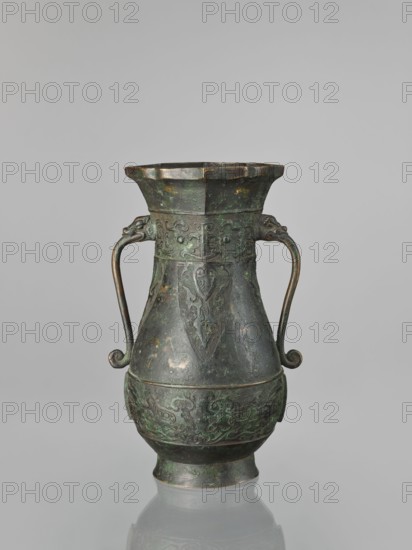Hu-type vase, hu vessel, Chinese, Ming dynasty 1368–1644, Xuande period 1426–1435, possibly backdated signatures, taotie ornamentation, ceremonial/ritual vessel, height 31.5 cm, width 17.0 cm., A tall metal vessel with a flared rim and bulbous body stands against a plain light gray background; the vessel shows a greenish patina with areas of dark brown and black, and a few lighter green and coppery spots. It has a pair of curved handles attached near the neck, each ending in a curled tip and featuring small sculpted animal head terminals, and the body is decorated with raised, carved motifs in low relief including stylized animal and geometric patterns around the midsection and a shieldlike decorative panel on the upper body. The surface appears aged and slightly mottled, and the vessel sits on a short foot that reflects faintly on the smooth surface beneath it.