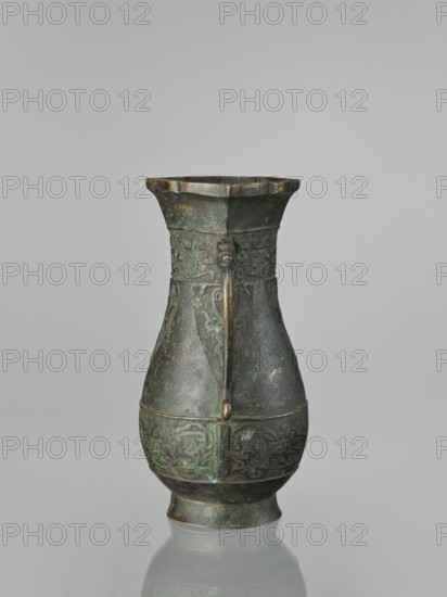 Hu-type vase, hu vessel, Chinese, Ming dynasty 1368–1644, Xuande period 1426–1435 — possible backdated signatures, taotie ornamentation, ceremonial/ritual vessel, height 31.5 cm, width 17.0 cm., An aged metal vessel stands upright against a neutral gray background, showing a patinated surface with green and dark bronze tones and touches of brown; it has a flared rim with a scalloped edge, a narrow neck that widens into a bulbous midsection, and a rounded foot; decorative relief patterns encircle the upper and lower sections, and a vertical strap-like handle runs from near the rim down the body, attached with a small ornamental element where it meets the midband; the surface shows areas of uneven coloration and subtle texture variations.
