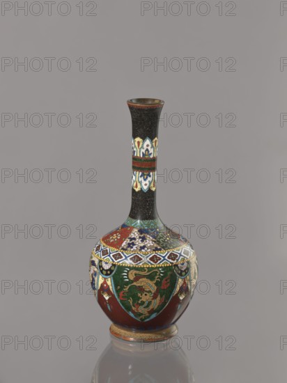 Vase, Japanese, Meiji 1868–1912, cloisonné enamel vessel decorated with birds, dragons, and phoenixes, ca. 1900, Japan; height 19.3 cm, width 8.9 cm, A tall, narrow-necked vase with a bulbous body stands against a plain gray background, showing a predominantly dark color palette with visible colors including black, deep red, green, white, blue, yellow, and gold; the neck is mostly black with a decorative band of lighter colors and a thin red rim, while the body features ornate multicolored patterns and motifs including green shields or panels with a golden dragon-like figure, red sections, floral designs in blue and white, diamond-shaped bands, and gold accents, and the vase sits on a small circular base that reflects faintly on the surface below.