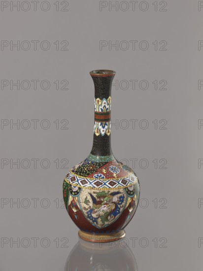 Vase, Japanese, Meiji 1868–1912, ca. 1900. Cloisonné enamel vessel decorated with birds, dragons, and phoenixes. Japan. Height 19.3 cm; width 8.9 cm., A tall, narrow-necked vase with a bulbous base sits against a plain gray background; the vase features a dark top section and neck with a textured black pattern, a band of decorative cream, blue, green, and red motifs on the neck, and a wider lower body decorated with intricate patterned panels in red, blue, green, cream, gold, and black, including floral and stylized bird-like designs, geometric borders, and scalloped medallions, with a small stepped foot rim in gold and brown and a faint reflection of the vase visible below.