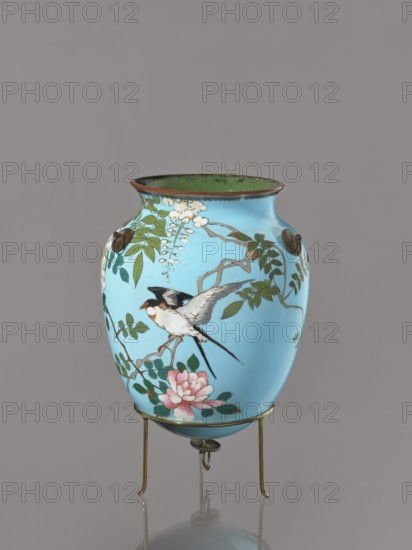 Vase, Japanese, Meiji–Taisho period ca. 1910–1920, cloisonné enamel, kachôga bird-and-flower imagery; decorated with flowers, wisteria, roses, and swallows; vessel, height 20 cm, diameter 15.3 cm., A rounded vase with a flared rim sits on a simple metal stand; the vase surface is a bright turquoise blue with painted natural motifs including a bird with black, white, and gray plumage perched among green leafy branches, white hanging blossoms, and a pink flower near the base; the rim interior appears green and the rim edge shows a brownish tone; the metal stand and small decorative fittings are brass-colored, and the background is a neutral gray.