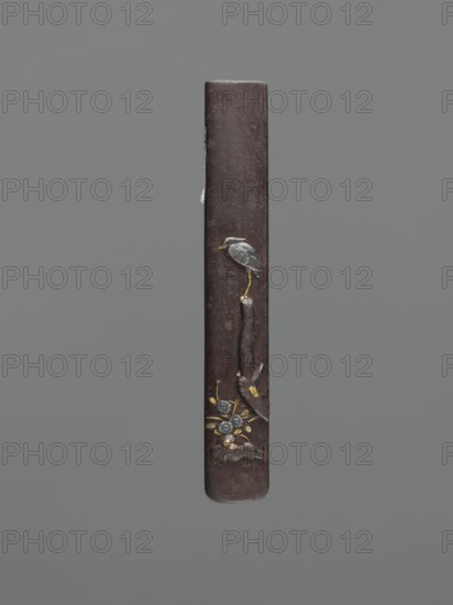 Knife handle kozuka, ; motif: herons; Japanese, Edo period 1603–1868, 18th century; materials/function: kozuka for a katana knife/edged weapon; dimensions: height 9.4 cm, width 1.4 cm, depth 0.6 cm., A slender vertical object with a dark brown, slightly textured surface against a gray background, featuring a small bird figure in gray and muted blue perched on a thin golden leg near the upper middle, a vertical dark brown element extending downward from the bird, and a cluster of decorative floral motifs near the lower section in metallic gold and silvery gray with touches of darker brown.
