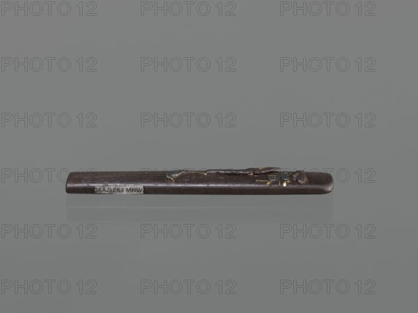 Knife handle kozuka, kozuka decorated with herons; Japanese, Edo period 18th century, 1603–1868; materials for sword/knife katana accessory; function: weapon component; dimensions: height 9.4 cm, width 1.4 cm, depth 0.6 cm., A narrow, elongated dark brown object lies horizontally against a smooth medium-gray background, with a glossy surface and a subtle reflection beneath it; toward one end there is a small raised decorative element that appears metallic with hints of gold and muted silver tones, and a few tiny areas showing reddish-brown and black details.