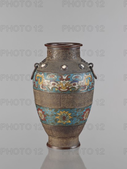 Vase, Chinese, Qing 1644–1911, 18th century, porcelain vessel decorated with lotuses, plant motifs, lotus tendril ornament, dragons and taotie iconography; height 30.5 cm; origin: China., An ornate vase stands against a plain light gray background, with a rounded body tapering to a narrower base and a slightly flared rim; the vase shows a mix of metallic brown areas with detailed etched patterns and colorful enamel bands in turquoise, yellow, red, blue, white, and green, featuring stylized floral and abstract motifs, with a few rounded metal studs near the shoulder and two dark metal loop handles on opposite sides; the surface alternates between textured bronze-toned sections and brightly painted decorative bands.