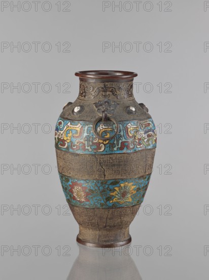 Vase, Chinese, Qing dynasty 1644–1911, 18th century, porcelain vessel decorated with lotuses, plant motifs, lotus tendril ornament, dragons and taotie iconography; height 30.5 cm; origin: China., A single ornate vase stands against a plain light gray background, with a bulbous body tapering toward a narrow base and a flared rim. The rim and base appear dark brown, while the main body shows alternating decorative horizontal bands: a few textured bands in a muted bronze-brown tone and several colorful bands featuring turquoise-blue fields with stylized floral and abstract motifs in yellow, red, green, and white. Near the neck there are small raised circular elements and metal ring handles attached to sculpted mounts in a darker metallic brown. The surface shows fine incised patterns and areas of subtle wear, and the vase casts a faint reflection on the glossy surface beneath it.